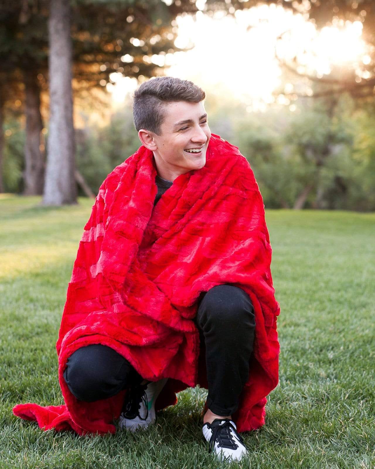 Boy crouched down at a park with a red minky blanket around him.