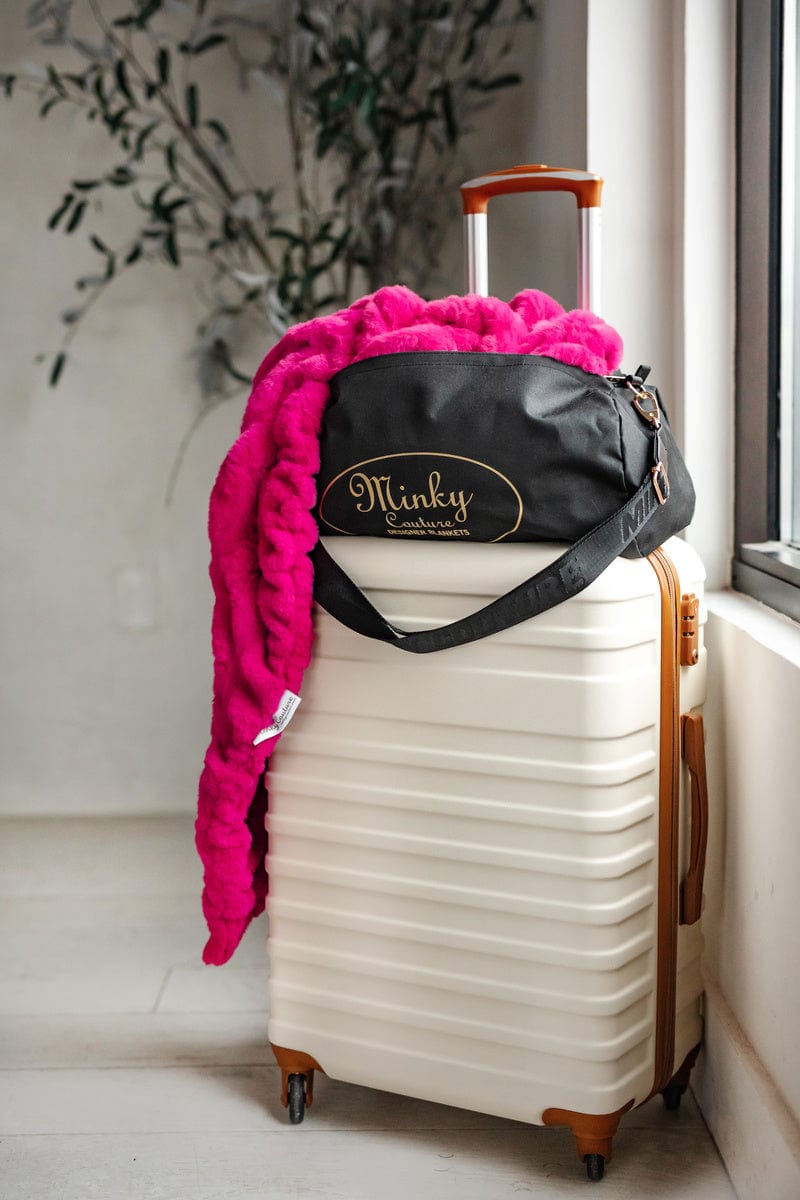 Bright pink blanket hanging out of a Minky Couture blanket travel bag that is on top of a cream rolling suitcase.