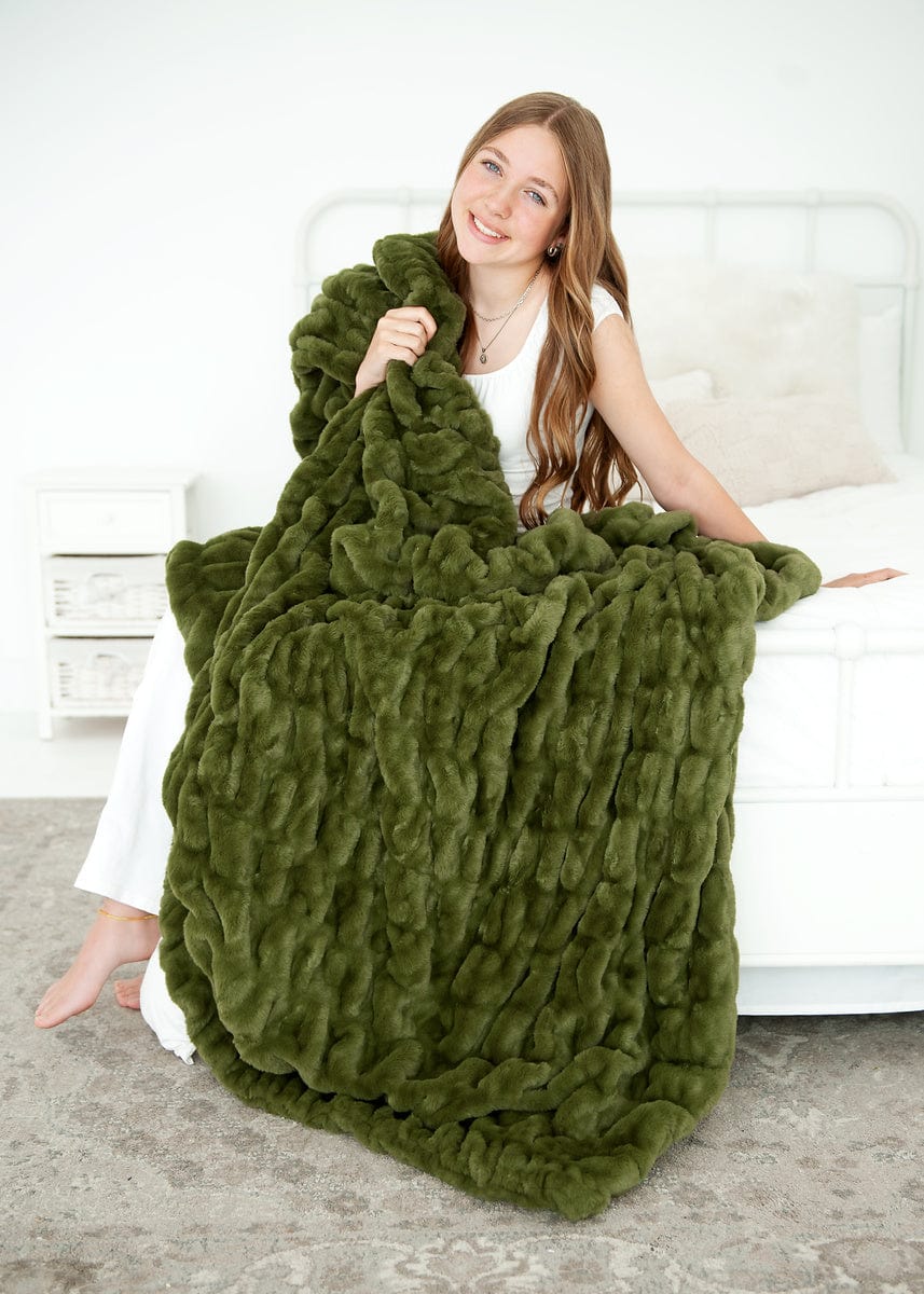 Young woman sitting on a bed wrapped up in a dark olive green plush blanket.
