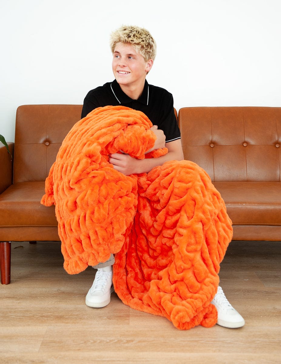 Young man sitting on a brown couch holding a vibrant orange blanket.