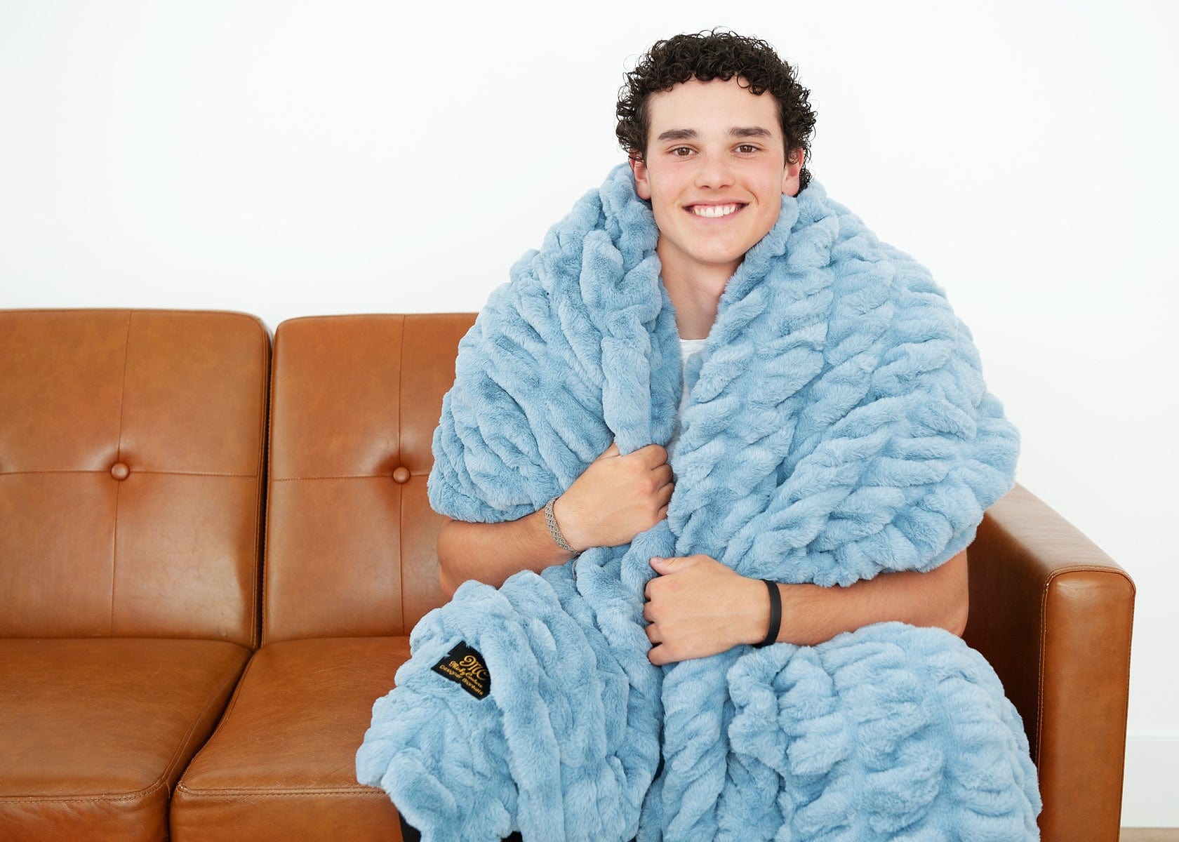 Young man sitting on a couch with a light blue ruched blanket around his shoulders.
