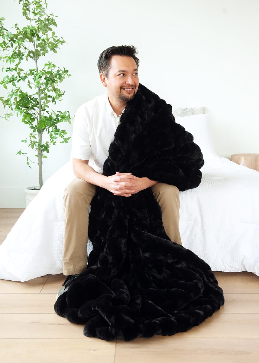 Man sitting on a white bed with a black blanket hanging off of one shoulder.