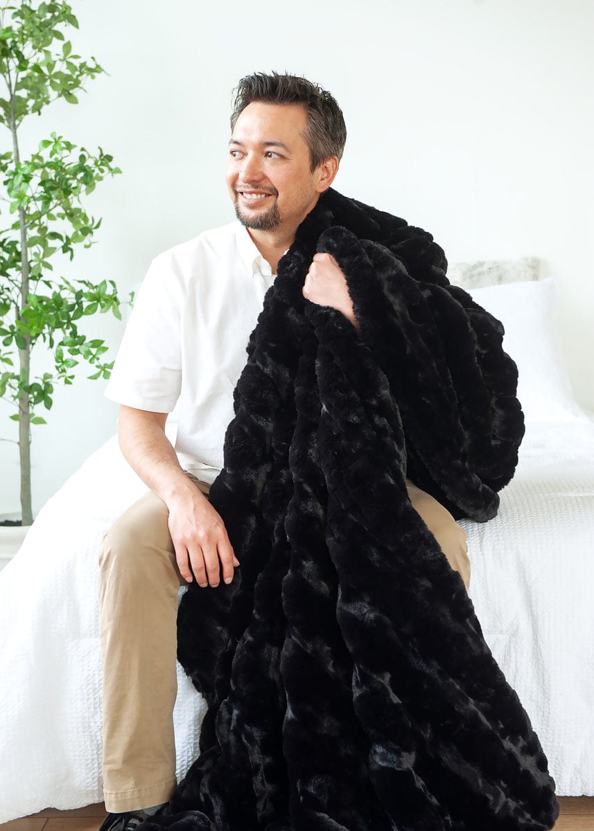 Man sitting on a white bed with a black blanket hanging off of one shoulder.