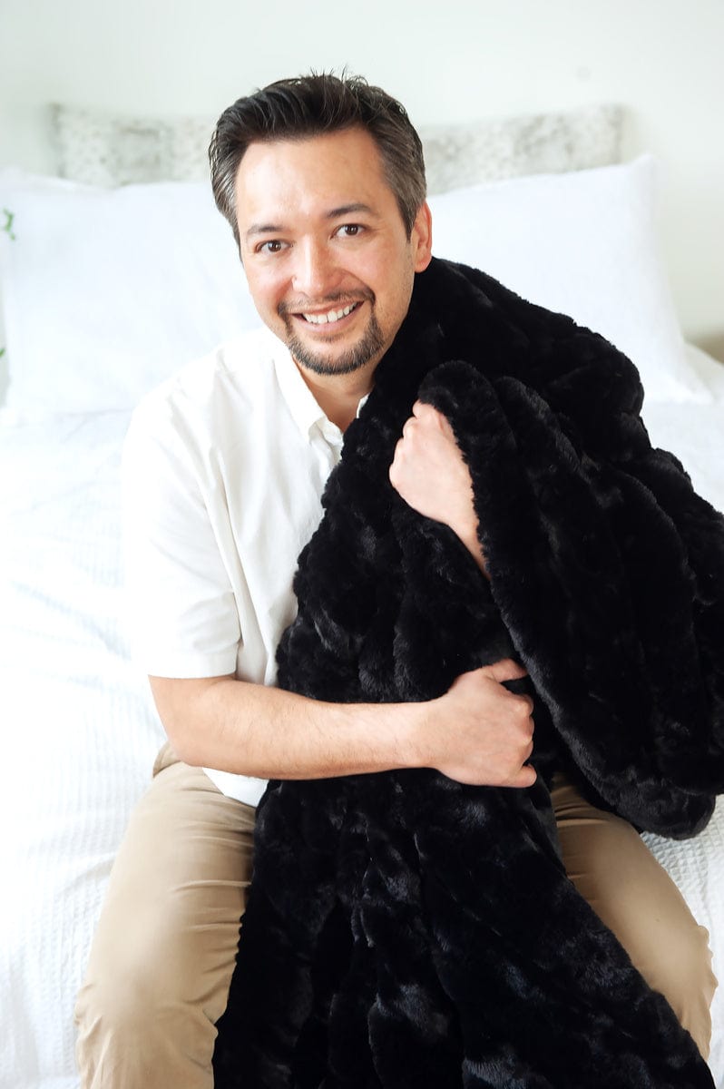Man sitting on a white bed while wrapped up in a black blanket.