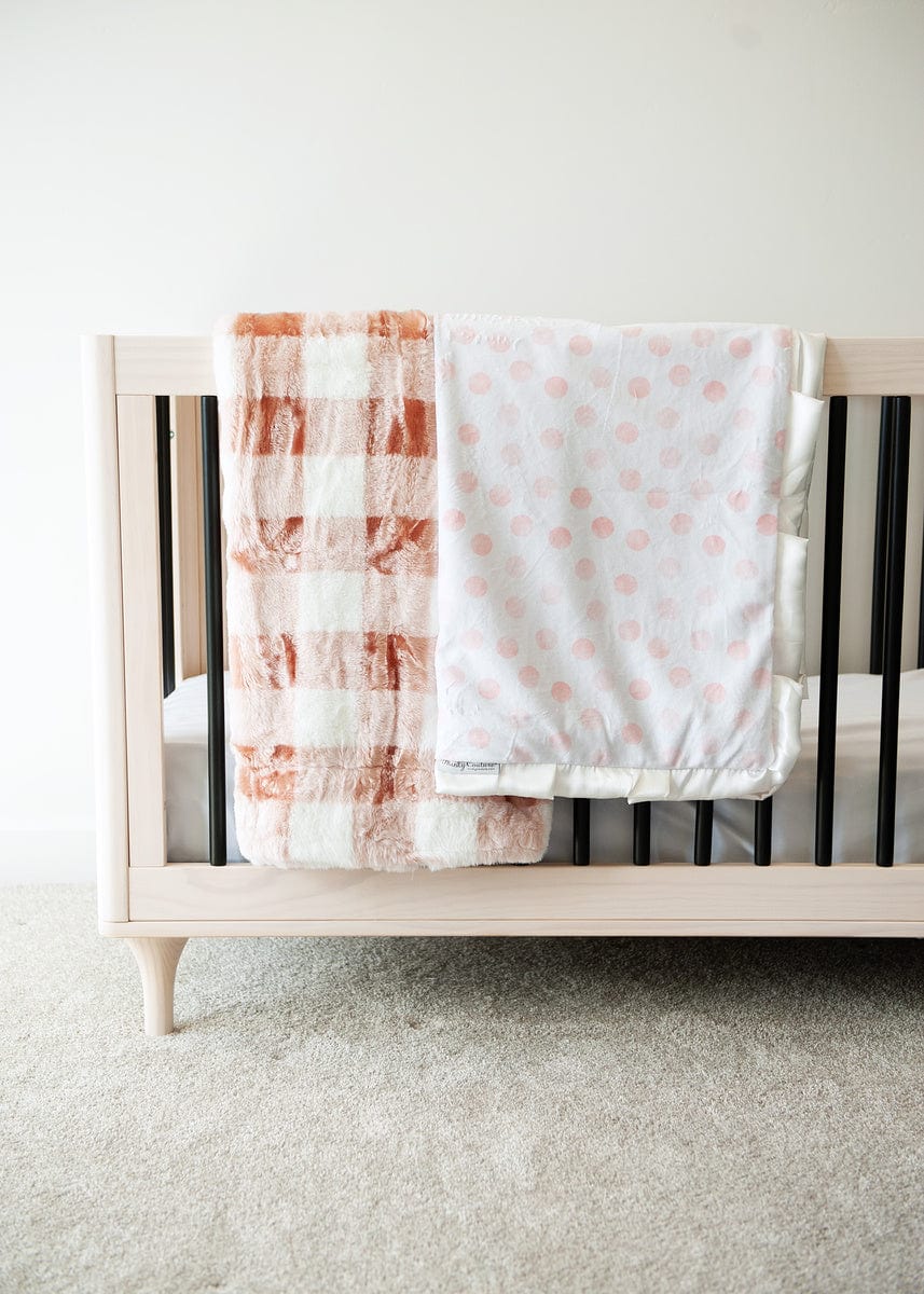 Blush and cream buffalo plaid plush blanket and white blanket with pink polka dots hanging on the side of a crib.