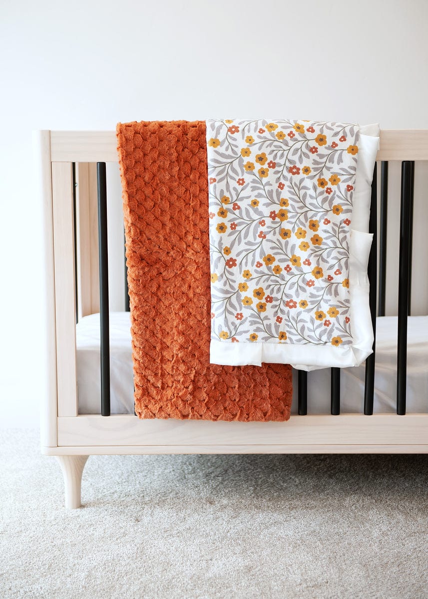 Waffle textured copper blanket and copper, mustard and gray floral blanket hanging over the side of a crib.