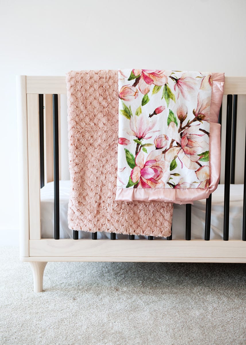 Blush waffle textured blanket and a pink magnolia patterned blanket hanging together over the side of a wooden crib.