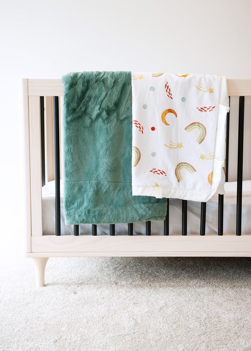 Thick mist green plush blanket and a rainbow, sun and moon themed blanket hanging over the edge of a crib.
