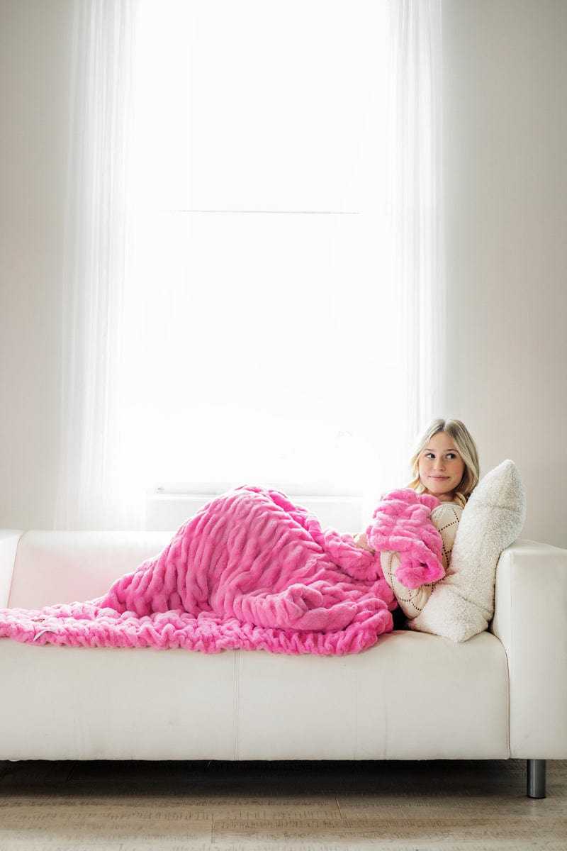 Young woman sitting on white couch snuggled up in a hot pink ruched plush blanket.