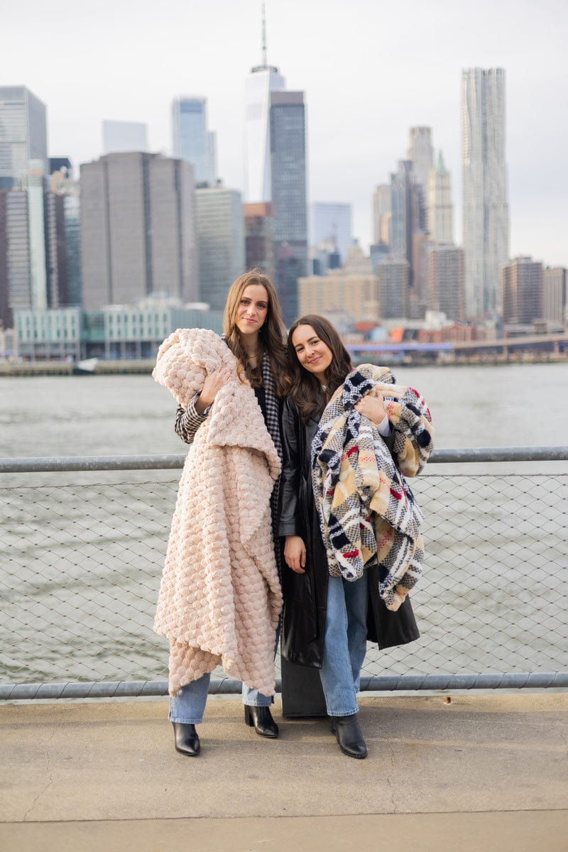 Two young women standing with a city in the background, each is holding a different minky couture blanket. 
