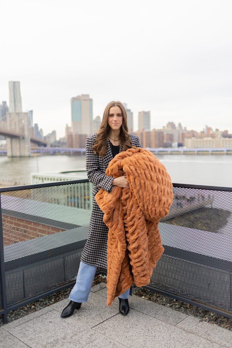 Young woman standing in a city holding a ruched copper plush blanket.