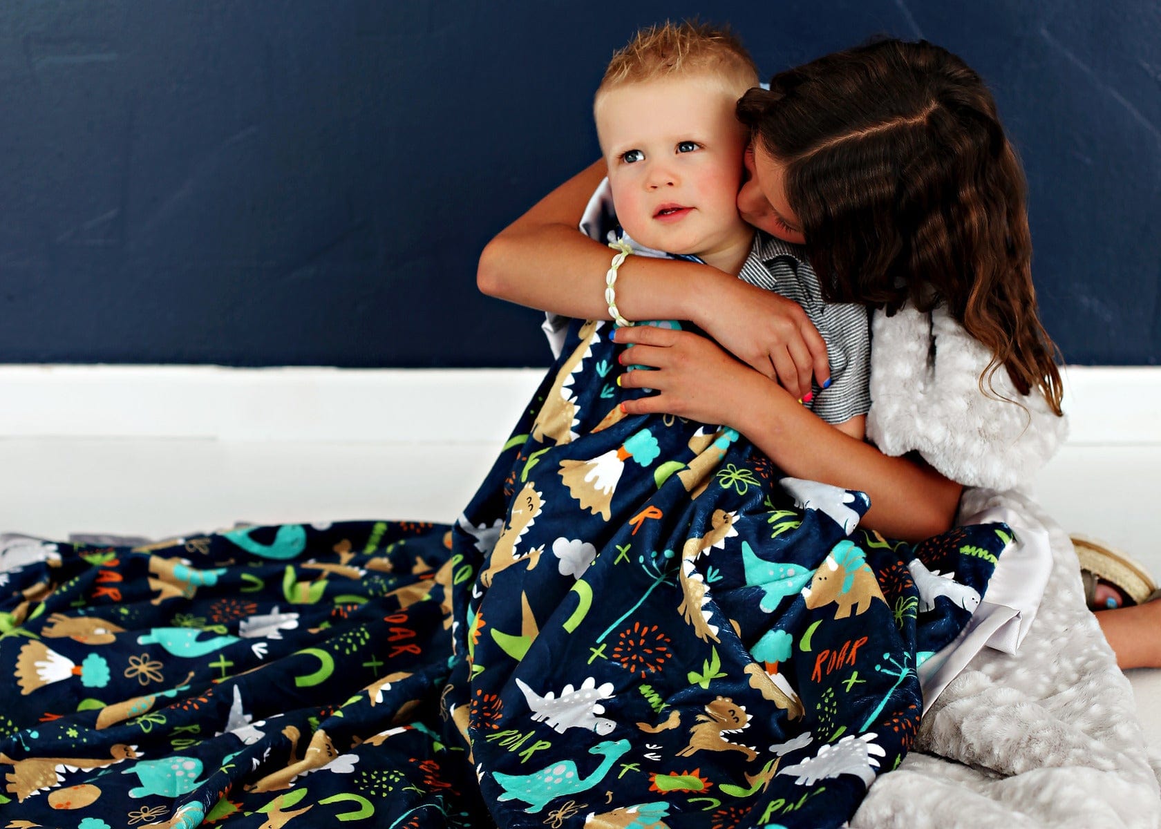 Sister kissing her little brother on the cheek and wrapping him up in a dinosaur themed blanket.