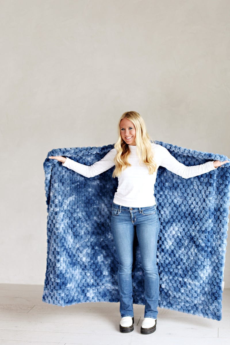 Young woman standing while holding a tie dye blue waffle textured blanket behind her back.