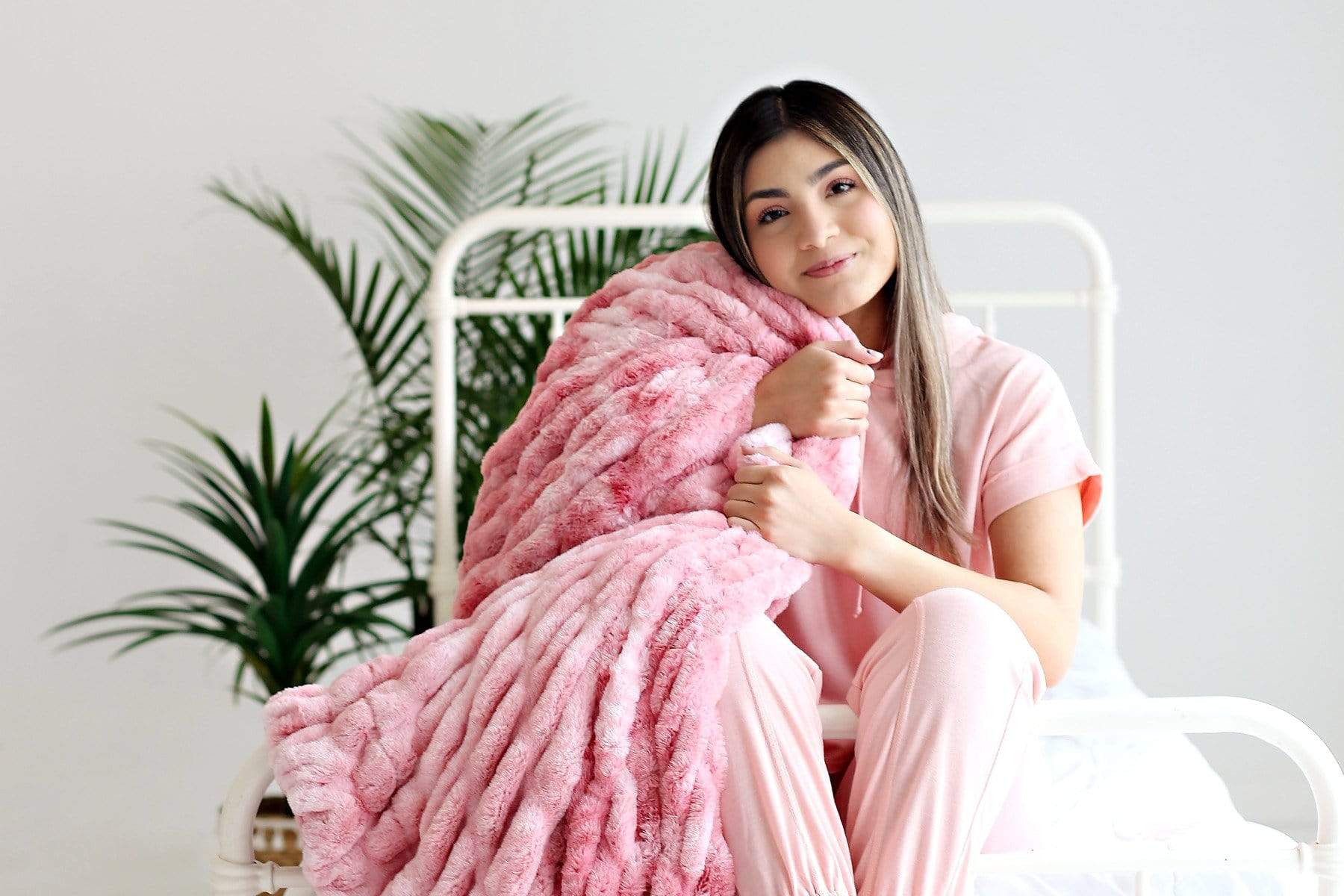 Young woman sitting on a bed with the cotton candy pink hugs stretchy blanket by Minky Couture over her shoulder. 