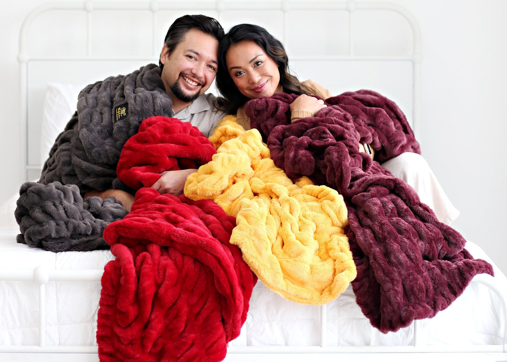 Couple sitting together on a white bed while wrapped up with four different colored stretchy blankets.