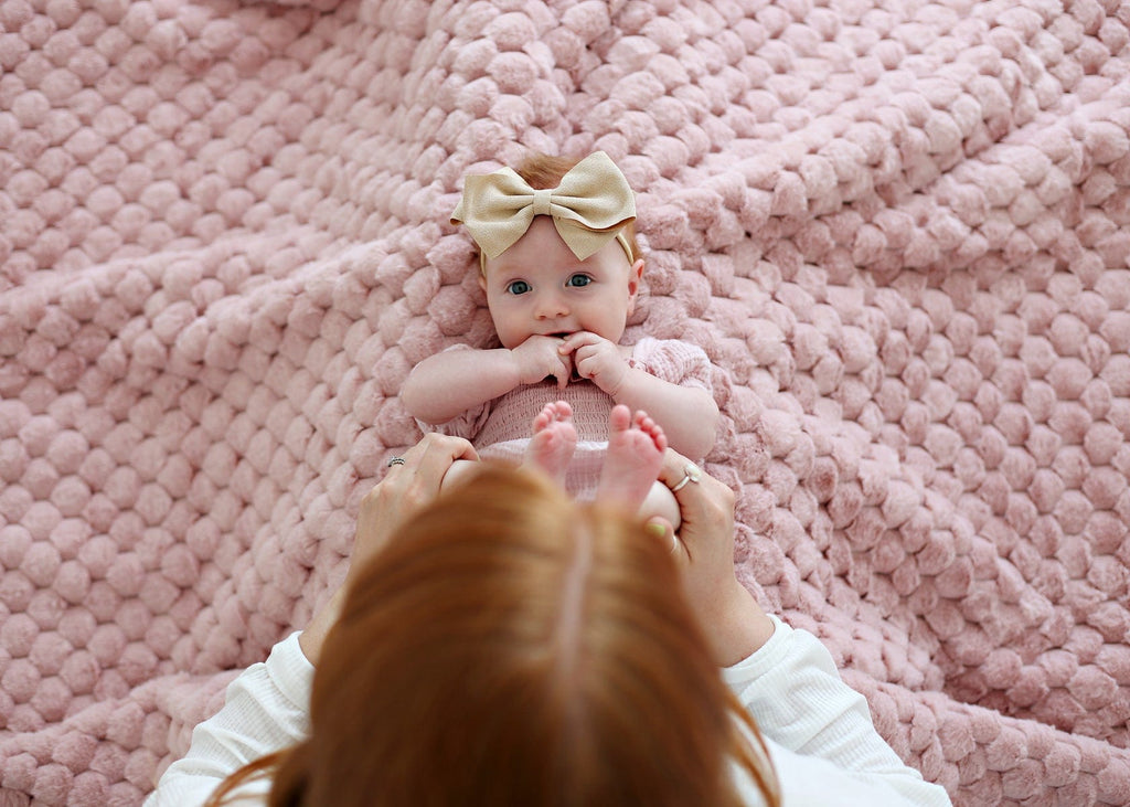 Mother playing with her young daughter while she lays on a soft blush colored waffle textured blanket.