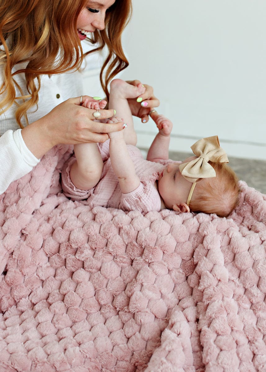 Mother playing with her young daughter while she lays on a soft blush colored waffle textured blanket.