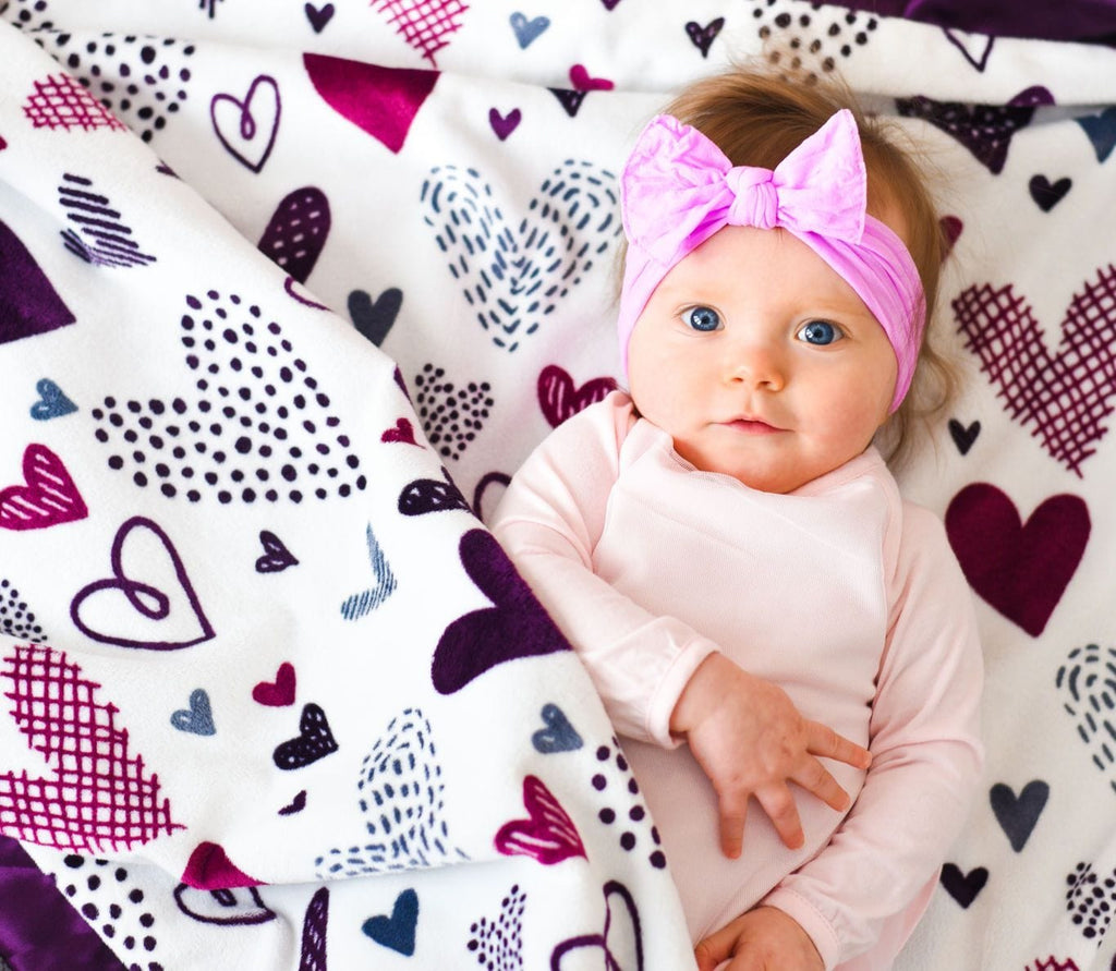 Baby girl cuddled in a Minky Couture designer blanket.  Blanket has purple hearts of all sizes on a cream background.
