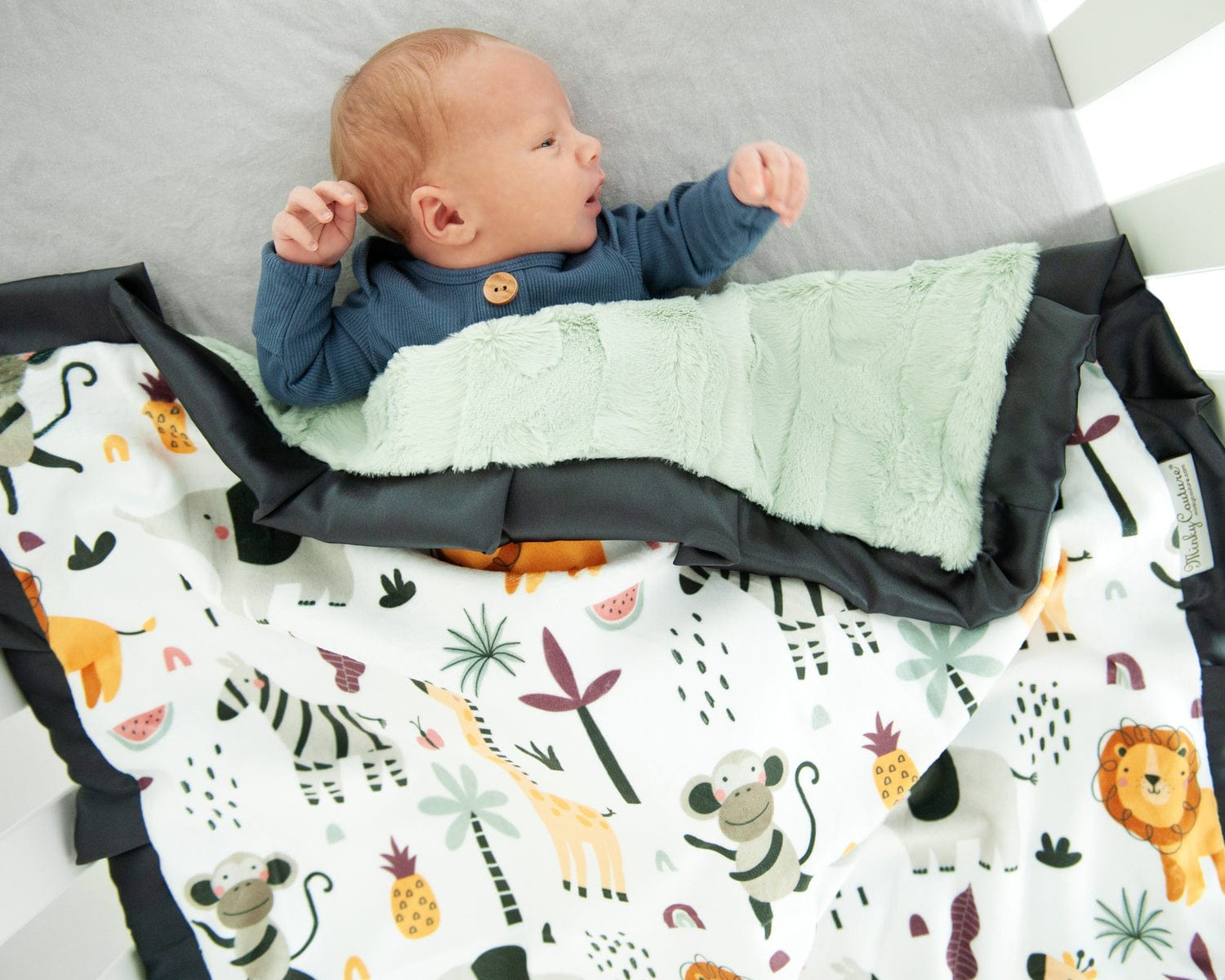 Baby boy laying in a crib with a cartoon zoo animal themed blanket. 