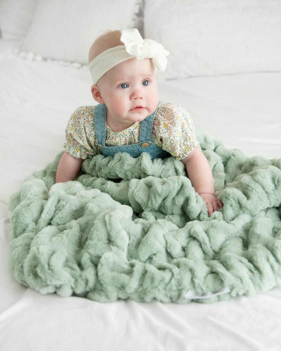 Baby girl sitting on a bed surrounded by a light green ruched blanket.