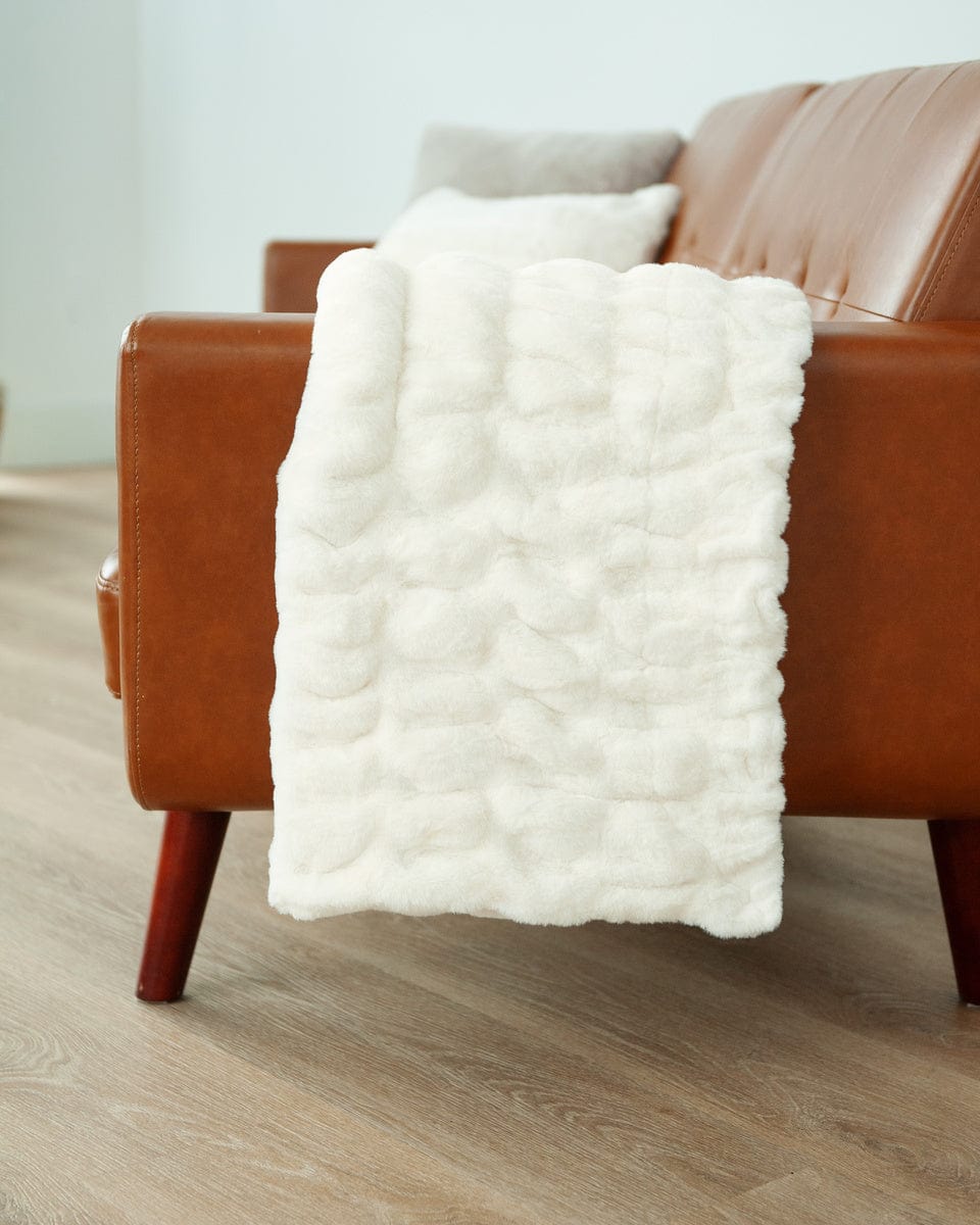 A cream plush blanket with ruched texture hanging over the arm of a brown leather couch.
