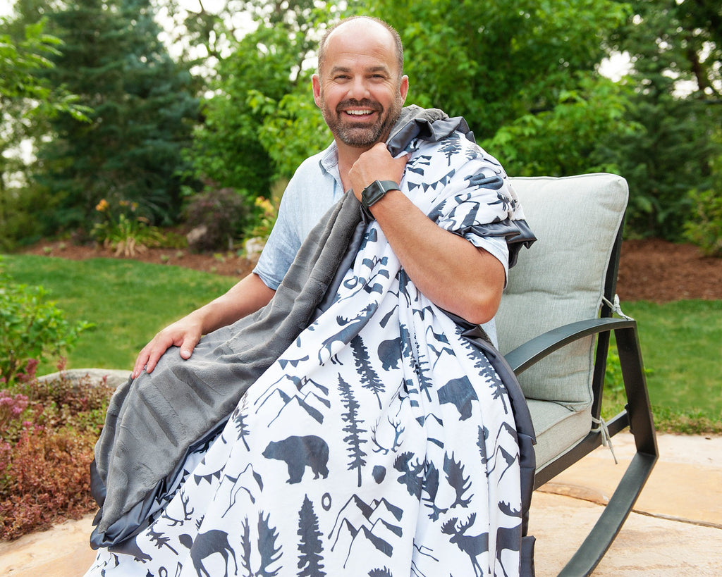 Man sitting outdoors holding a gray and white mountian themed blanket with mountains, antlers, bear, moose and more on it.
