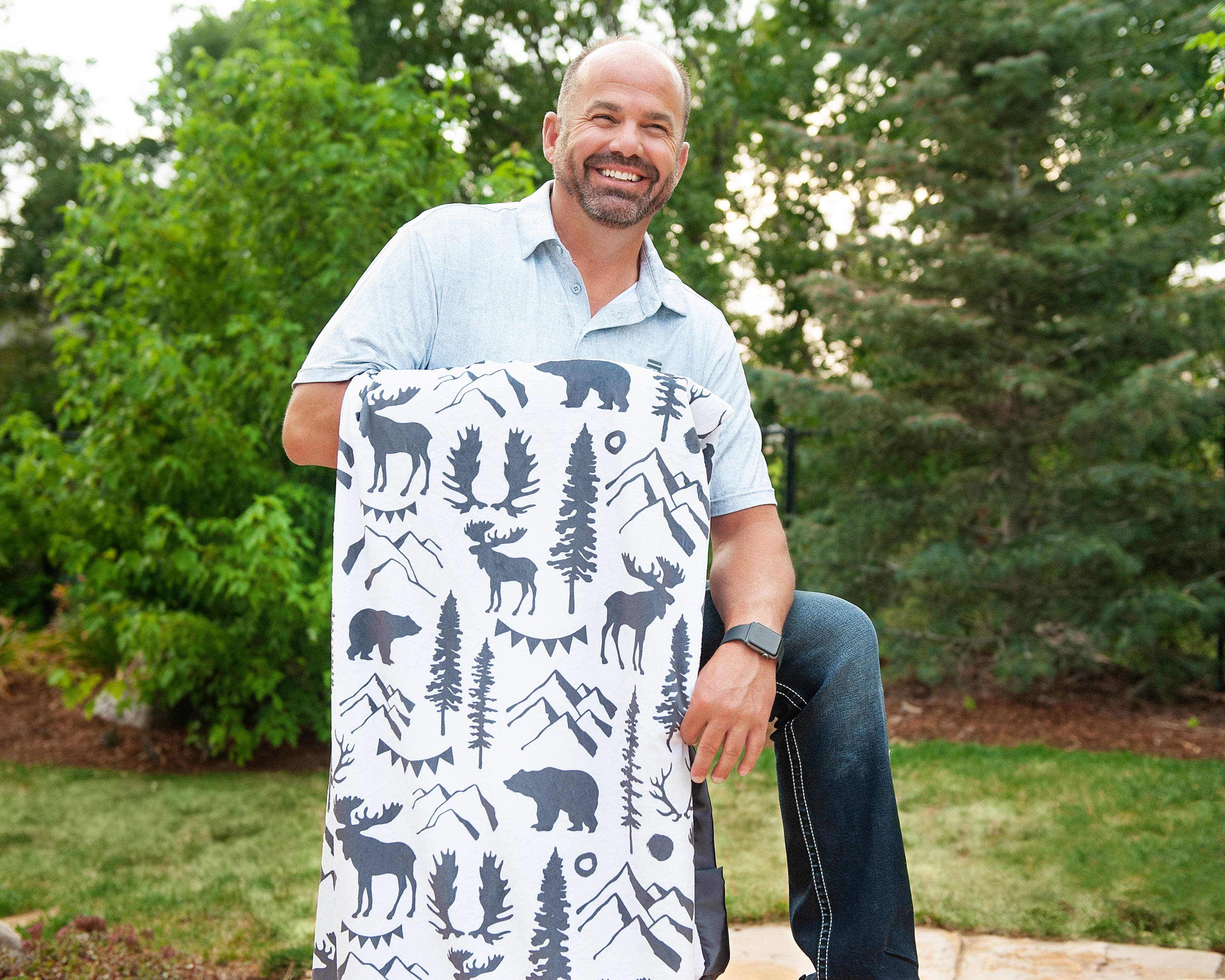 Man standing outdoors holding a gray and white mountian themed blanket with mountains, antlers, bear, moose and more on it. 