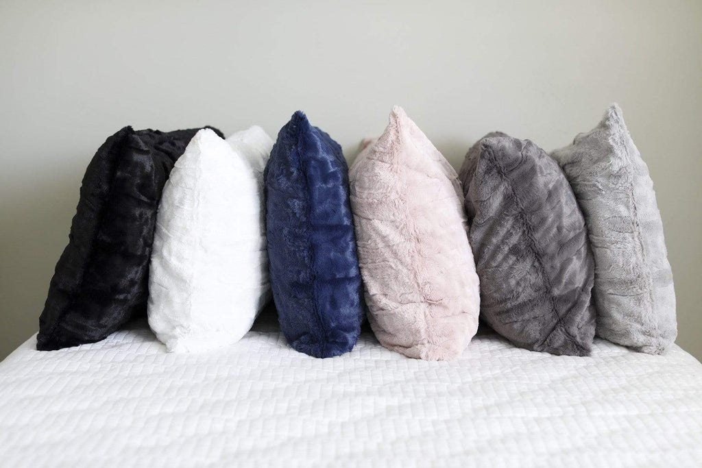 Six plush pillows by Minky Couture lined up on a bed. Colors shown from left to right are black, cream, navy, blush, charcoal and gray.