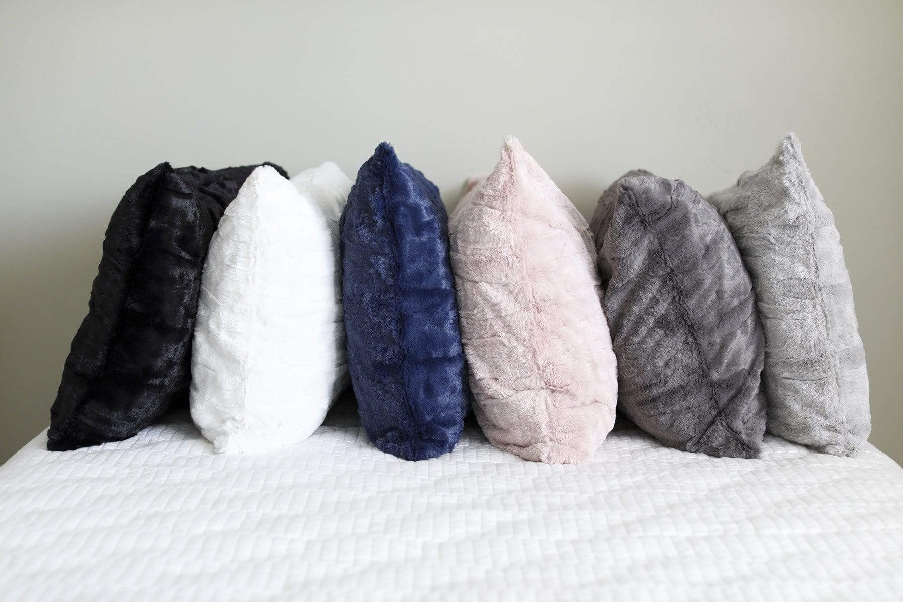 Six plush pillows by Minky Couture lined up on a bed. Colors shown from left to right are black, cream, navy, blush, charcoal and gray.