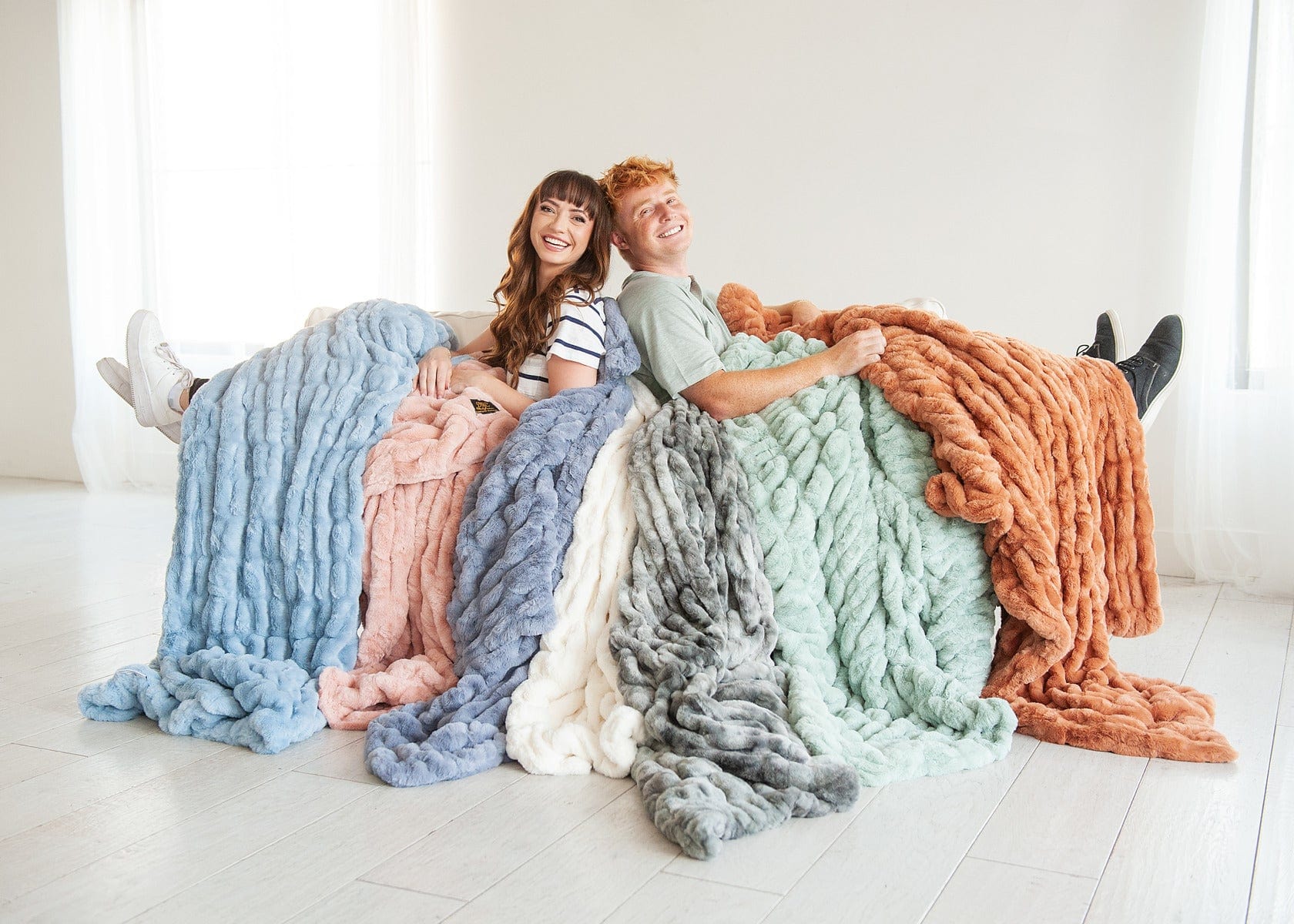Young woman and young man sitting back to back on a couch surrounded by seven different colored plush blankets.