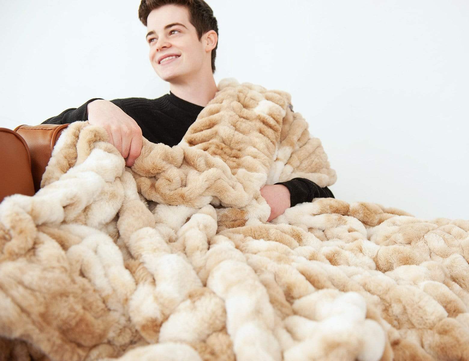 Young man sitting on a couch covered up with his Cotton Candy Caramel stretchy hugs blanket by Minky Couture. 