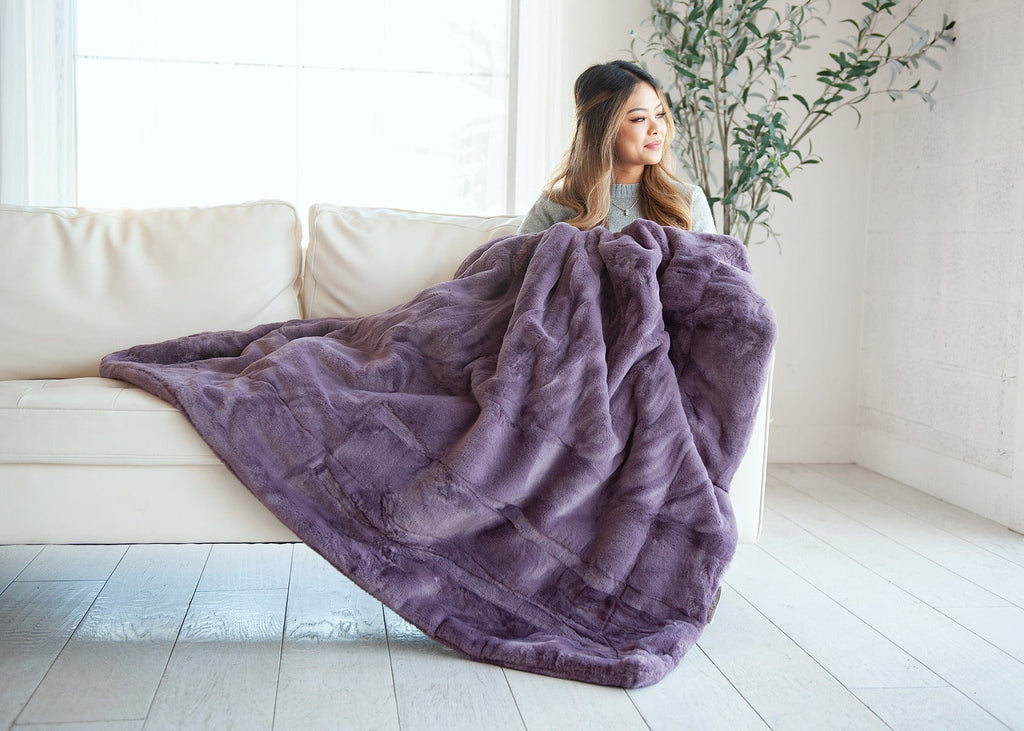 Young woman sitting on a couch holding a heather purple thick plush blanket.