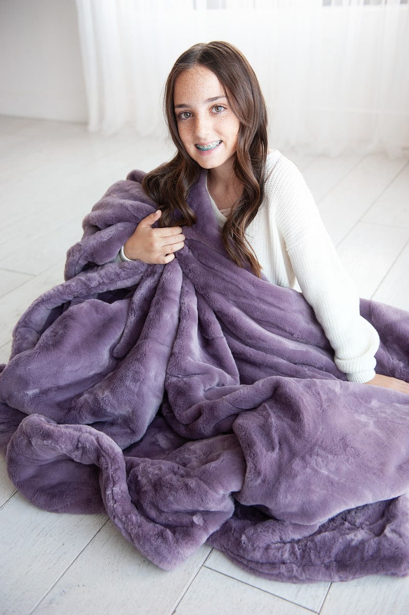 Girl sitting on the ground wrapped up in a heather purple thick plush blanket.
