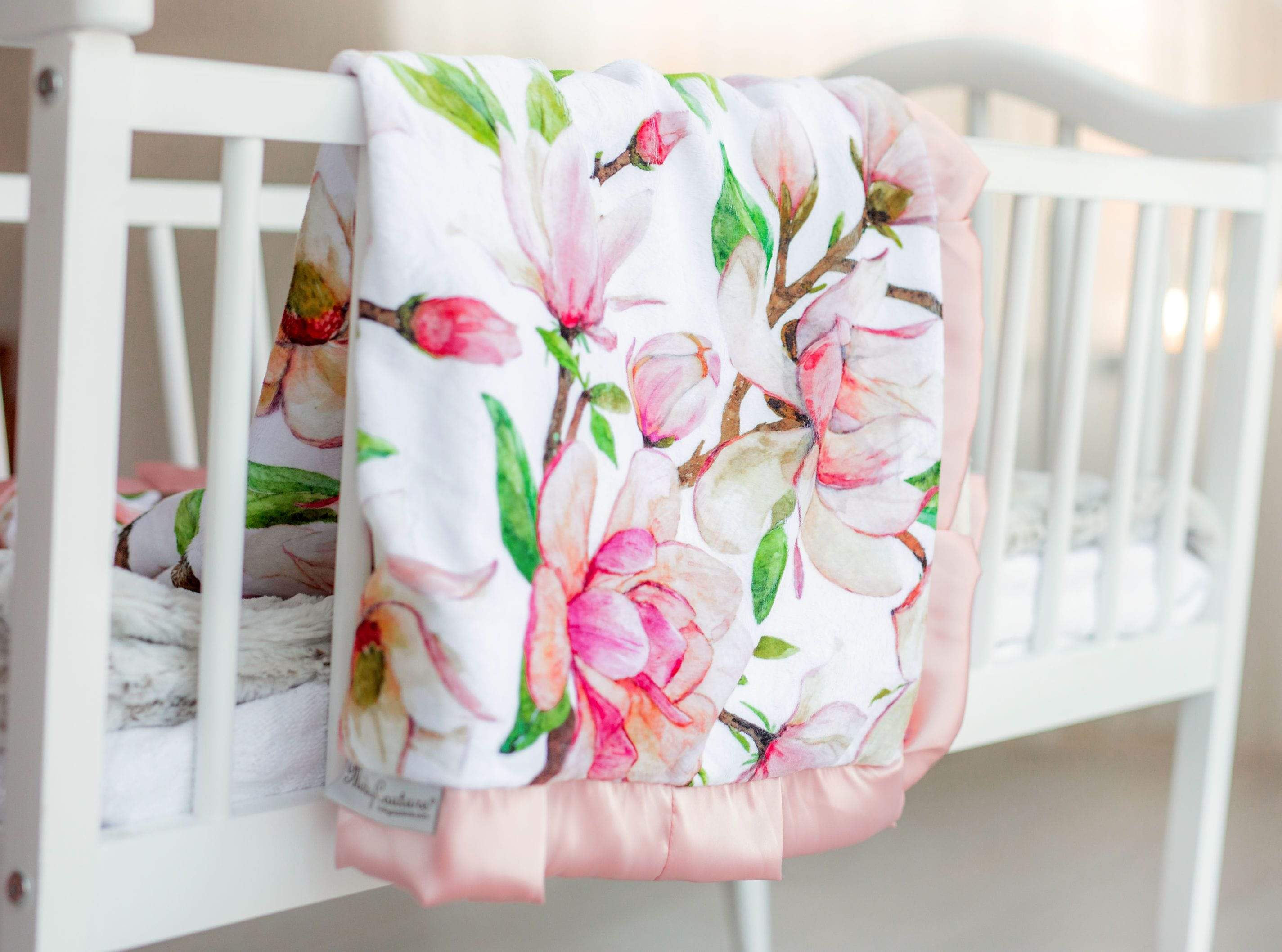Pink Aspirations blush and pink floral patterned blanket by Minky Couture laying over the side of a white crib.