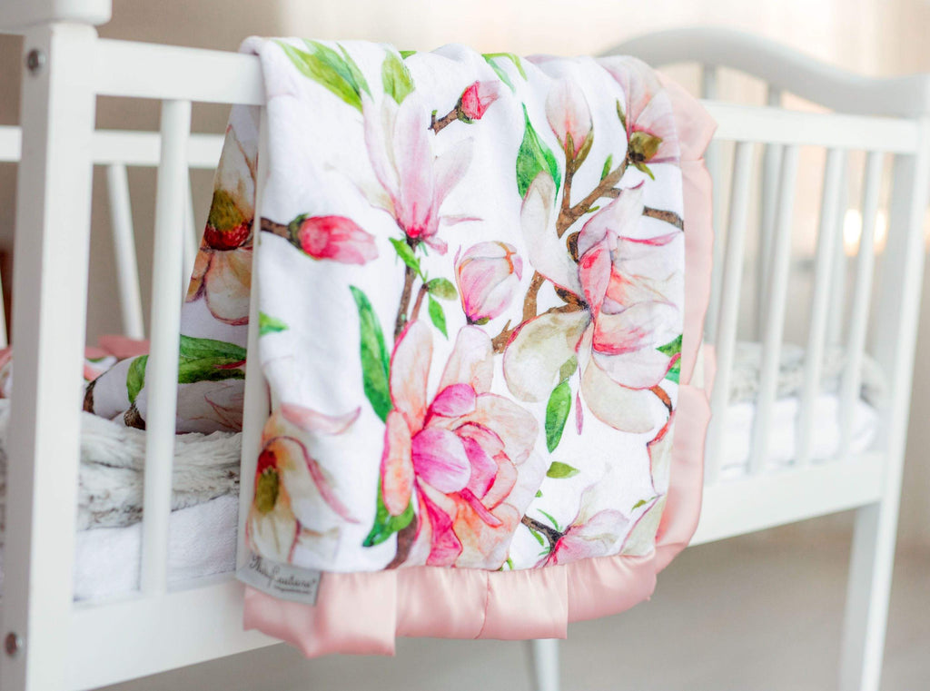 Pink Aspirations blush and pink floral patterned blanket by Minky Couture laying over the side of a white crib.