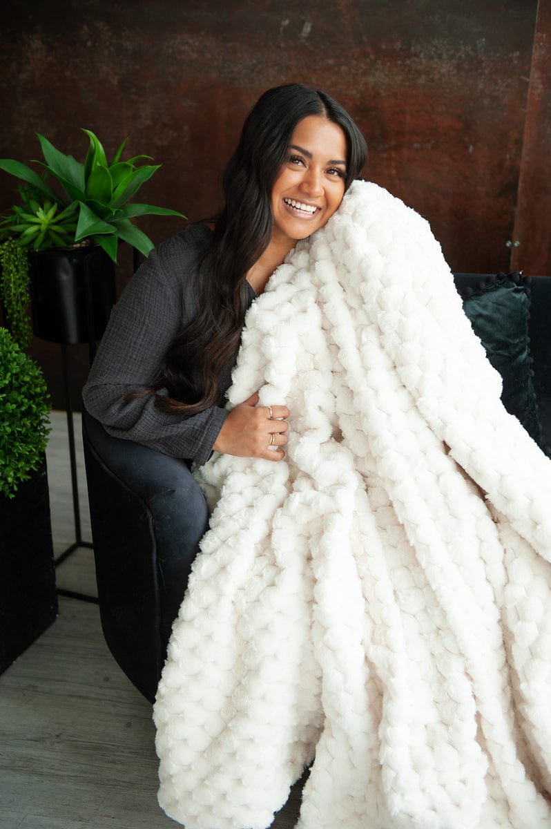 Young woman sitting on a couch holding a cream waffle textured blanket. 