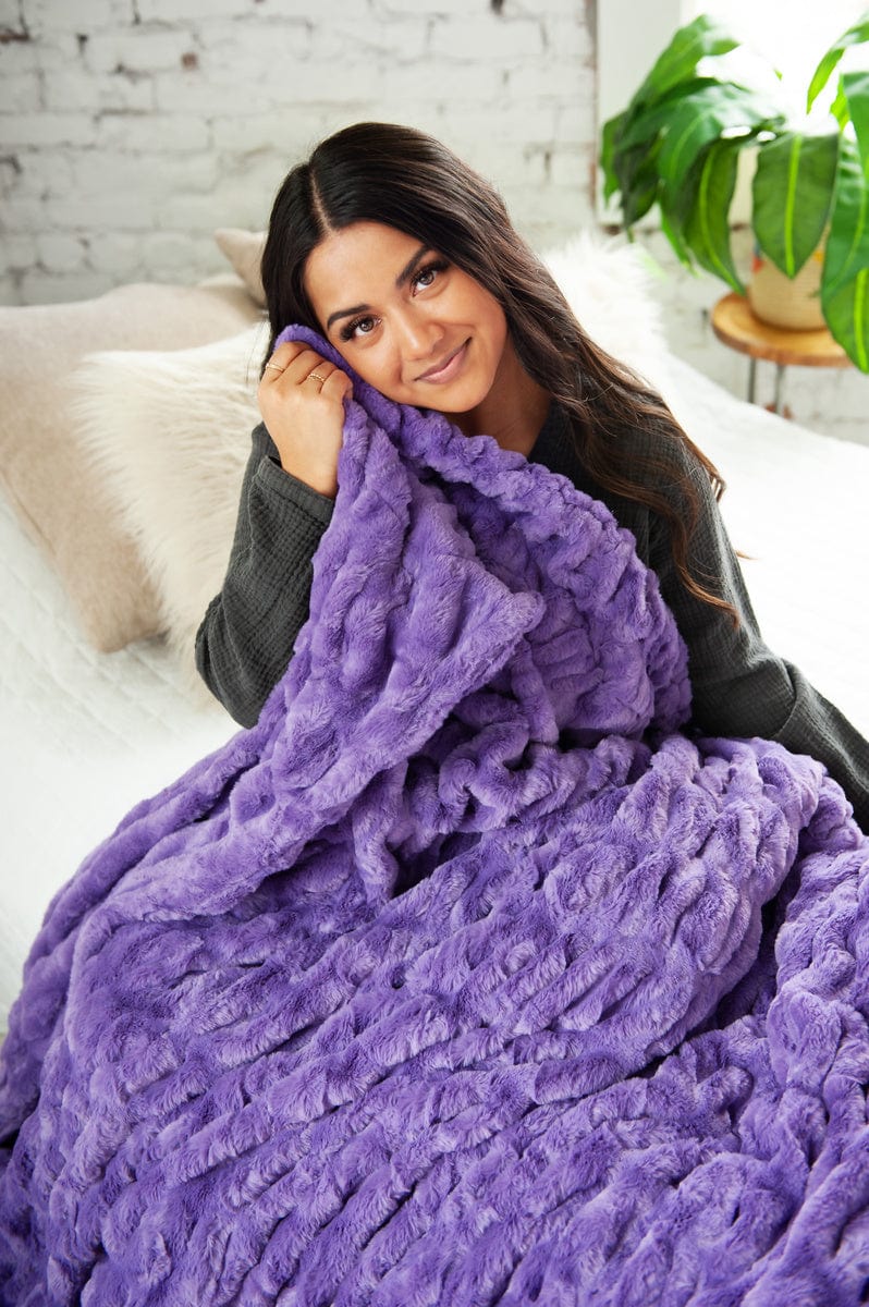 Young woman sitting on a bed with a vibrant purple ruched blanket held near her face. 
