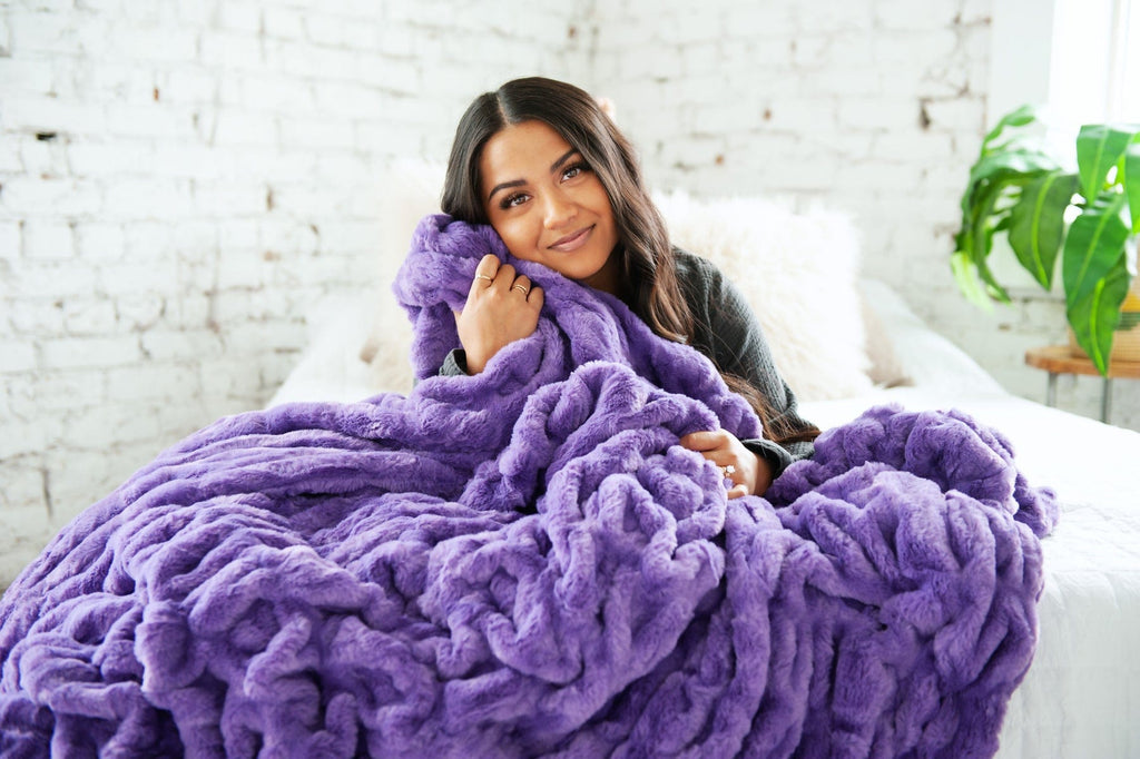 Woman laying on a bed with a vibrant purple ruched blanket in her arms.