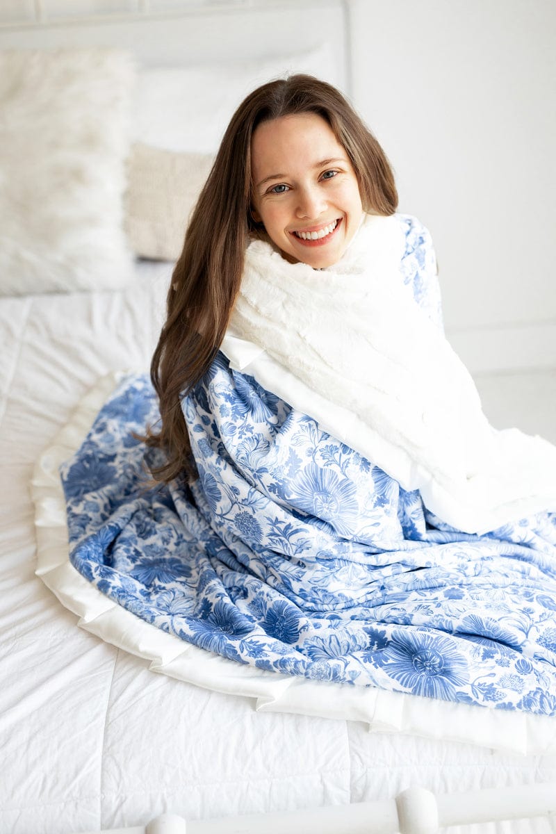 Young woman sitting at the edge of a bed with a blue and white floral blanket wrapped around her shoulders.