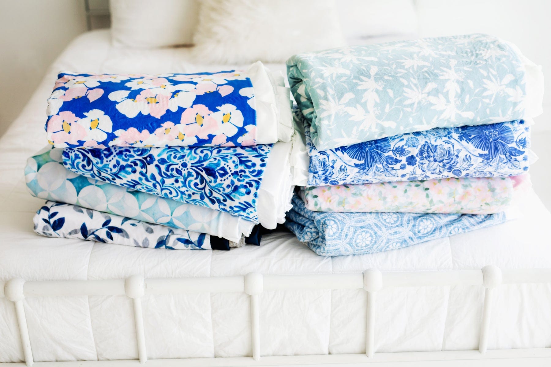 Eight different patterns of blue Minky Couture blankets folded and stacked at the end of a bed.