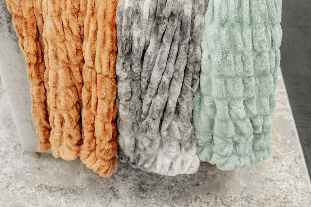 A copper, a variegated gray and a mist green colored ruched blanket hanging from the back of a couch.