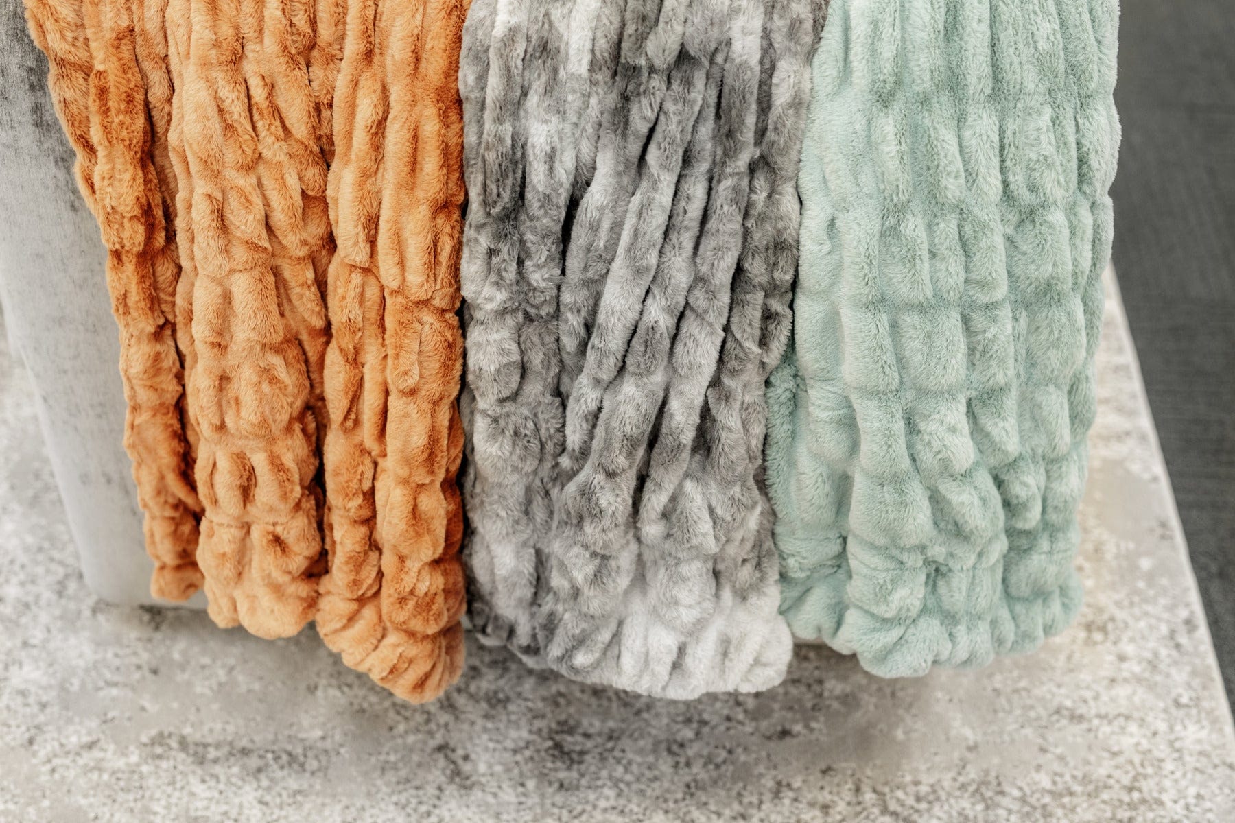 A copper, a variegated gray and a mist green colored ruched blanket hanging from the back of a couch.