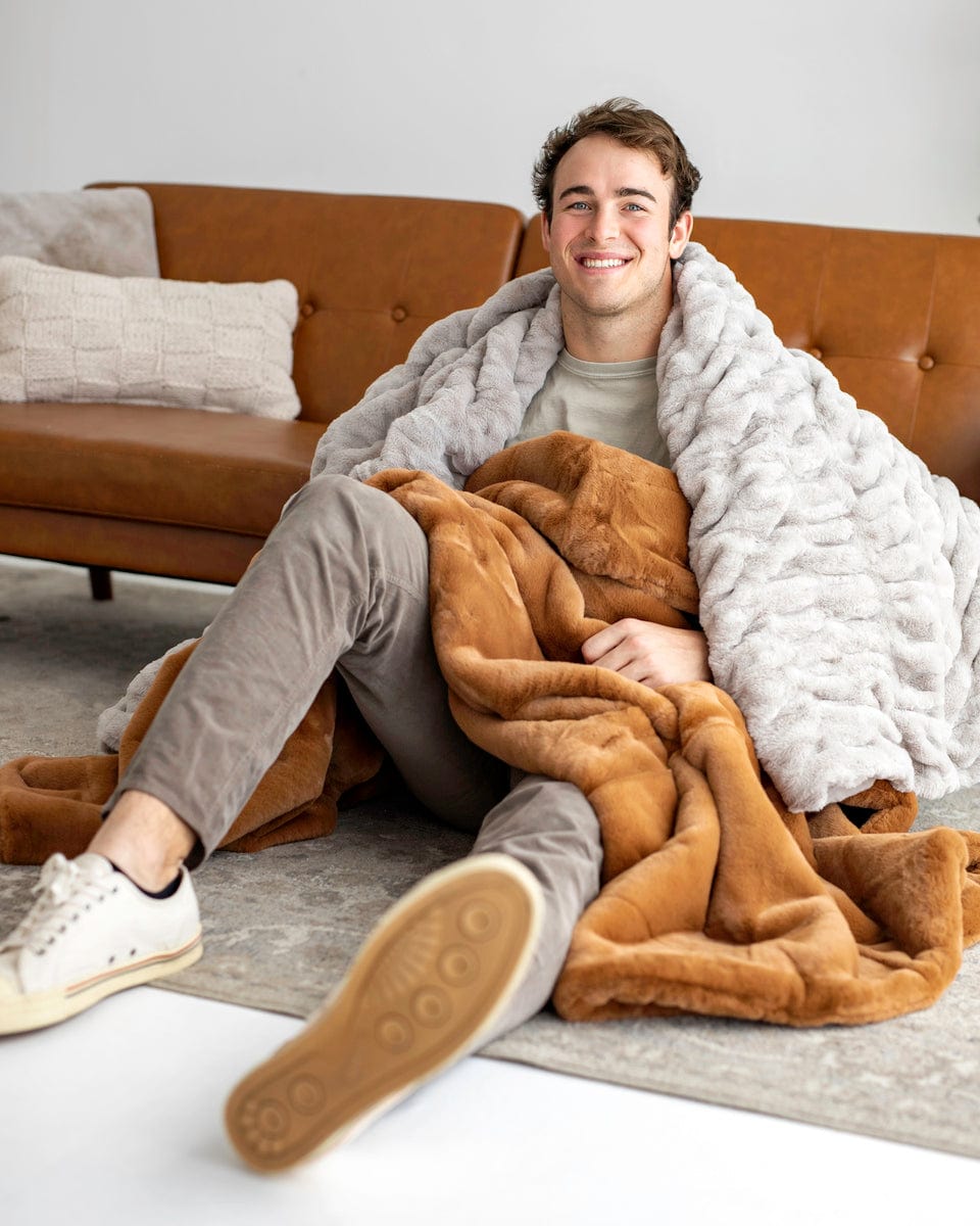 Young man sitting in front of a brown couch with a light gray blanket around his shoulder and a copper blanket over his legs.