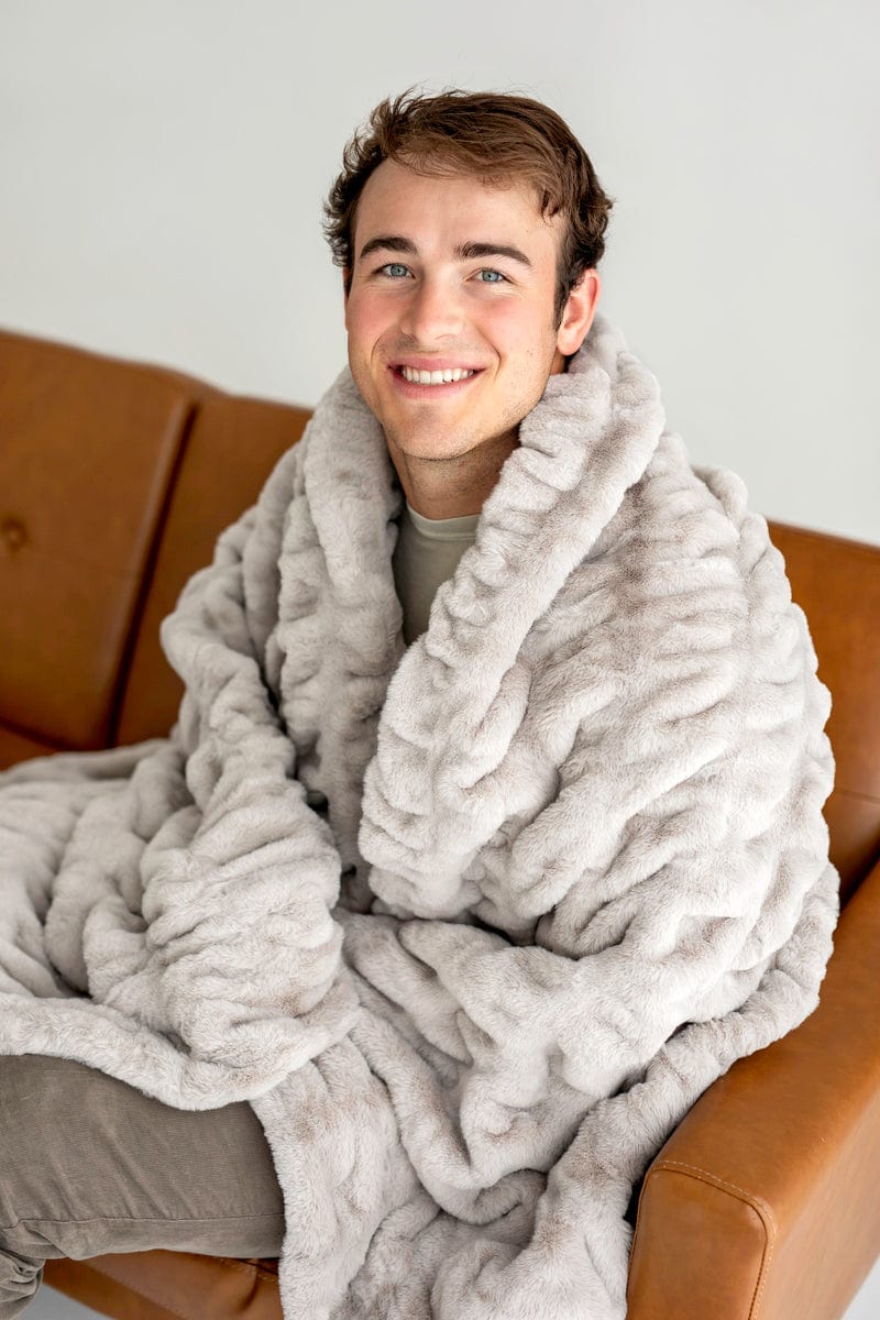 Young man sitting on a brown couch wrapped up in a light gray ruched blanket by Minky Couture.