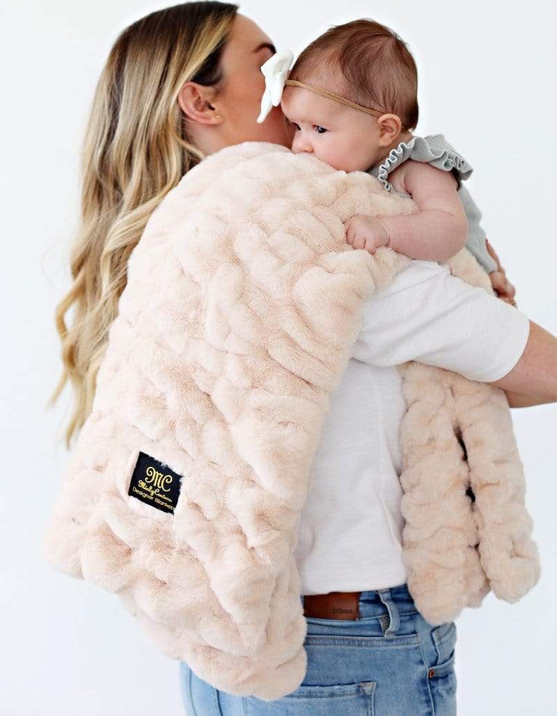 Mother holding her baby to her shoulder where a folded cream plush blanket lays.